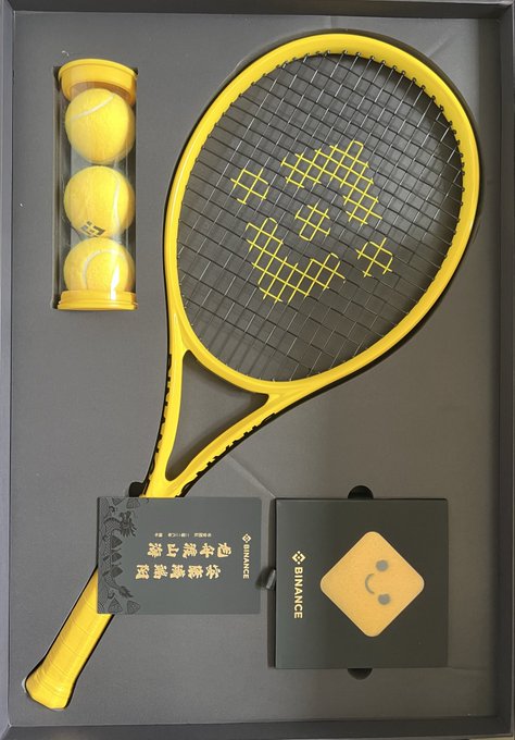 A yellow tennis racket with matching yellow balls in a clear tube, displayed in a black gift box alongside a small black cube with a smiley face logo and a SINIACE branded card.