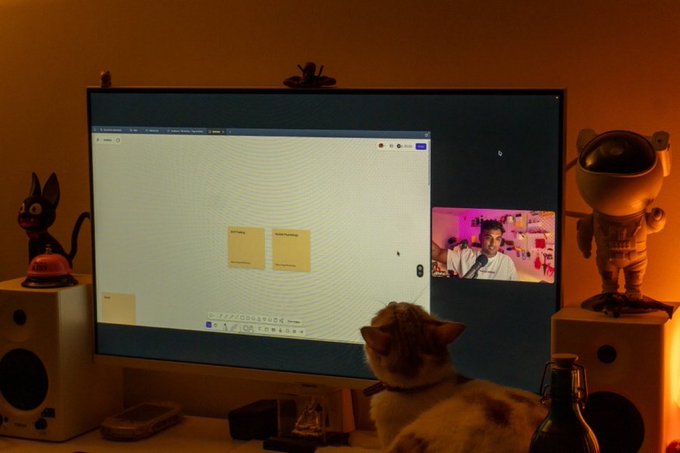 A cozy desk scene featuring a cat watching a video call on a monitor with sticky notes, flanked by an Astro Boy figurine, a small black cat figure, and warm ambient lighting.