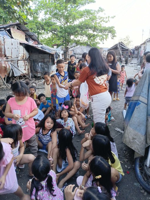 A large group of children and a few adults gather in what appears to be an impoverished neighborhood with makeshift shelters, as a woman in an orange shirt distributes items to the kids.