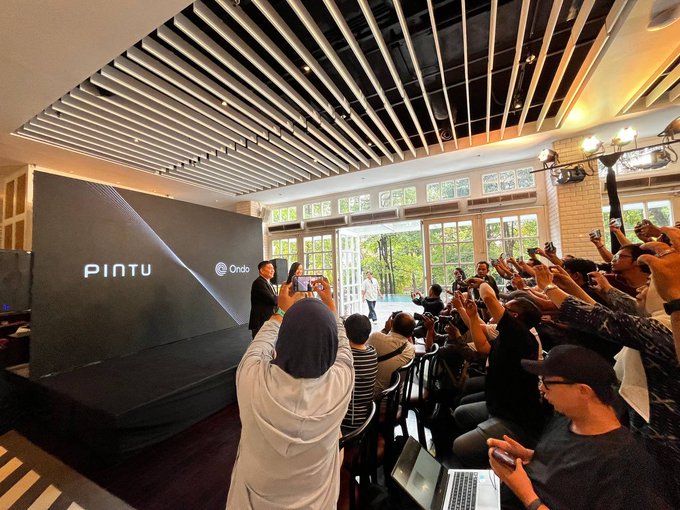 Crowded indoor event space with a black stage backdrop displaying 'PINTU' and 'Ondo' logos, where an audience is taking photos with their phones.