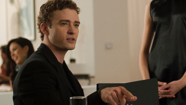 A curly-haired man in a black blazer sits at a table with a drink in front of him, looking toward a woman standing nearby in a black dress.