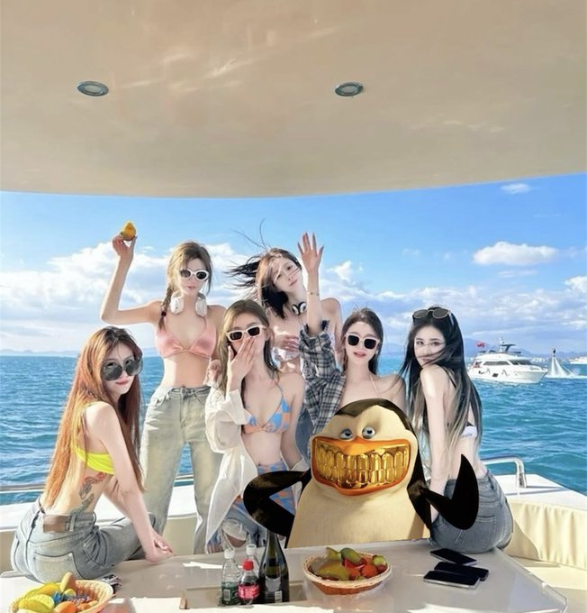 A group of young women in bikinis pose on a yacht with the sea and boats in the background, featuring a photoshopped character with a large grinning mouth in the center of the group.