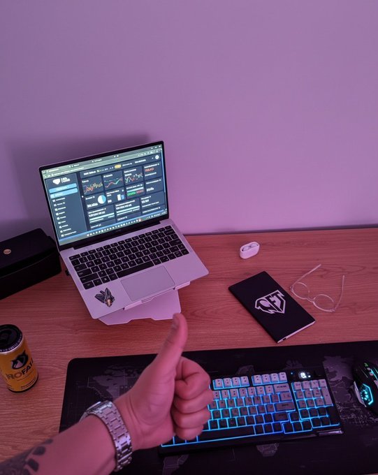A thumbs-up gesture in front of a desk setup featuring a laptop displaying analytics dashboards, a backlit mechanical keyboard, mouse, glasses, and a can of Tropik soda, with a notebook bearing a logo that appears to match "Quant." The image visually reinforces the "GM" (good morning) crypto greeting and the post's theme of trust and intelligence in AI, as the workspace setup reflects a tech-savvy trader engaging with Quant's data-driven platform.
