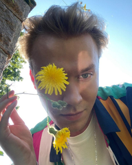 A close-up selfie of a young man outdoors with bright yellow dandelions held in front of his face.