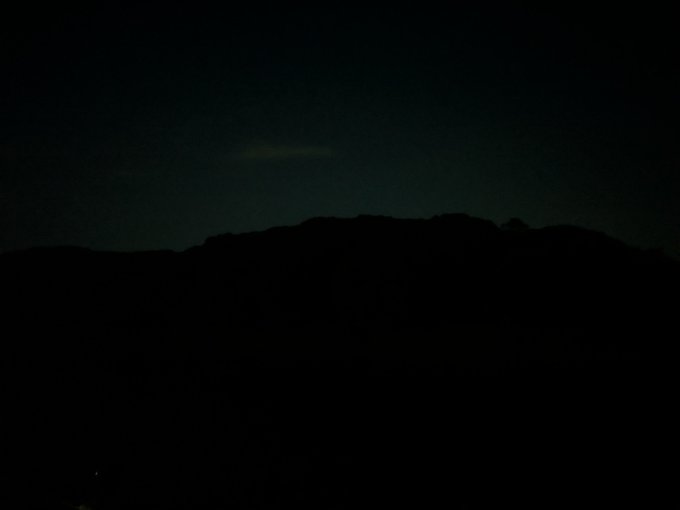 A very dark nighttime photograph showing the faint silhouette of a mountain ridge against a dim sky, with minimal visible detail.