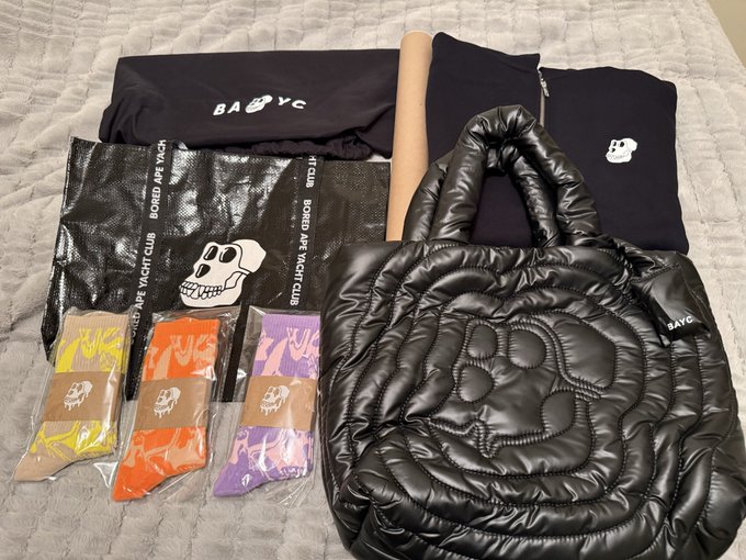 A flat lay of merchandise on a white bed including a black puffy "BAYC" rose-shaped bag, a black mesh tote, t-shirts, and three pairs of skull-branded socks in yellow, orange, and purple packaging.