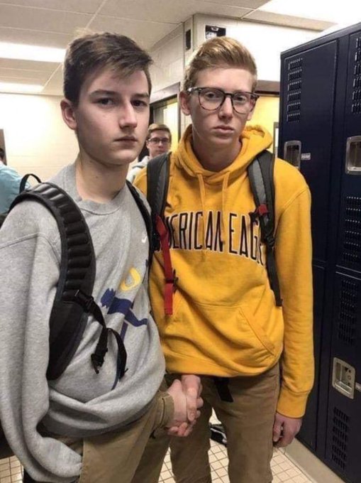 Two teenage boys pose together in what appears to be a school hallway with lockers; one wears a gray sweatshirt and backpack, while the other wears a yellow "American Eagle" hoodie and glasses.