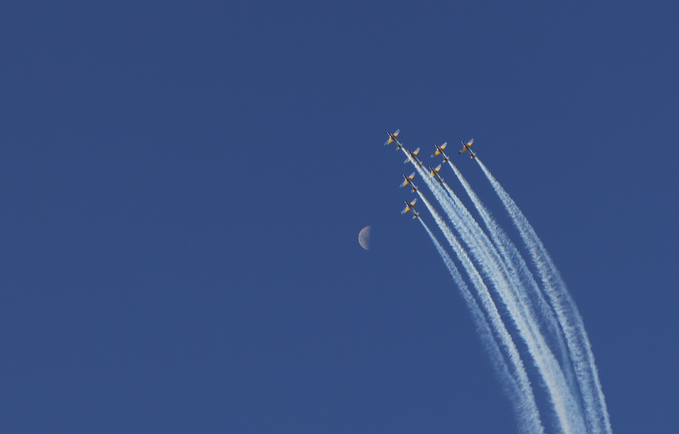 Five jets fly in formation against a clear deep blue sky, leaving long white contrails as they pass near a pale crescent moon.