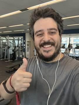 A smiling man gives a thumbs-up while wearing wired earphones in what appears to be a gym or office fitness area.