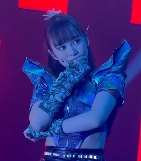A performer in a metallic blue and silver stage costume with spiked shoulders poses with their hand near their face under purple and blue stage lighting.