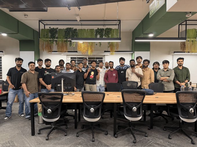 A team of around 20 young developers stands together in a modern office space with hanging plants and ergonomic workstations, celebrating the successful conclusion of Episode 1 of the Dodo Payments build IRL event, with several members wearing branded merchandise from the event.