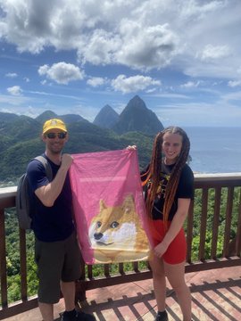 A man wearing a yellow hat and a woman stand together on a balcony holding up a pink towel featuring the Doge Shiba Inu meme, with tropical mountains visible in the background.