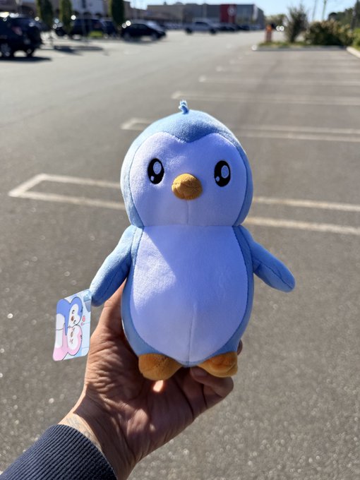 A hand holds a blue penguin plush toy outdoors in a parking lot, with a "BAB" tag attached to its arm.