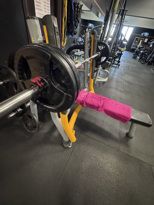 A Smith Machine barbell loaded with 20kg weight plates rests on a bench in a gym, with a bright pink towel draped across the seat.