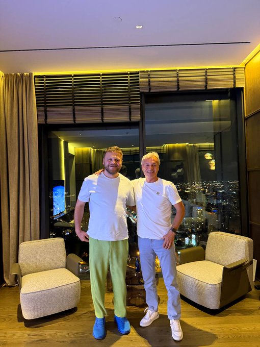 The image features two men standing next to each other in a room with large windows that provide an impressive view of the city skyline at night. The man on the left, wearing blue shoes, has his arm around the other man who is dressed in white. They are both smiling and appear to be enjoying their time together. In front of them, there are two chairs placed next to each other, adding a cozy touch to the scene.