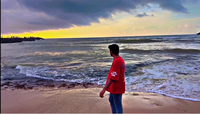 A man in a red t-shirt and jeans standing on a sandy beach looking out at ocean waves under a sunset sky.