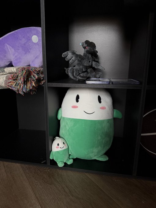 Two Cupsey plushies—a large green and white character with rosy cheeks and a smaller matching mini version—sit on a black shelf alongside a gray shark plush and a purple whale plush, perfectly matching the caption celebrating the crypto mascot in the creator's office.