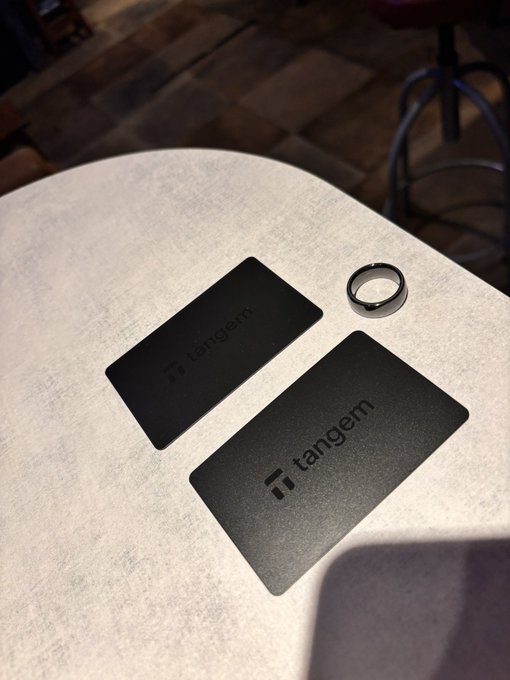 Two black 'tangem' branded card readers and a smart ring resting on a light-colored fabric table.