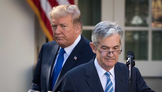 Two men in dark suits stand at a podium with a microphone, with an American flag visible in the background, illustrating the article's claim that President Donald Trump is demanding that Federal Reserve Chair Jerome Powell resign from the Fed board.