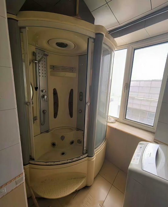 A bathroom interior featuring a corner shower cabin with multiple body jets and a showerhead, situated next to a large window, with a white washing machine visible in the lower right corner.
