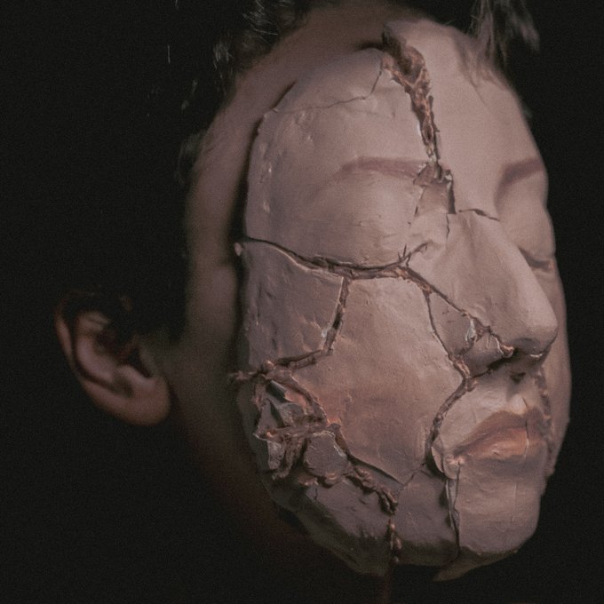 A close-up photograph of a person's face covered in a cracked, clay-like mask or prosthetic, showing detailed fissures and a reddish, flesh-toned appearance against a dark background.