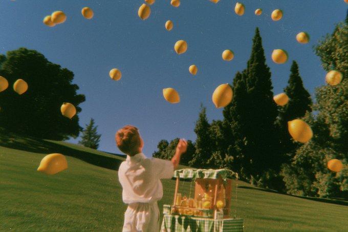 A surreal photograph showing a person in white standing in a grassy field, tossing yellow lemons into the air, with a lemon stand visible nearby.