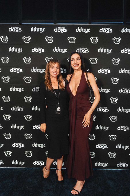 Two women pose together in front of a black step-and-repeat backdrop featuring 'digital dogs' and 'ddnyc' logos, with one woman wearing a black long-sleeved dress and the other wearing a burgundy plunge-neck gown.