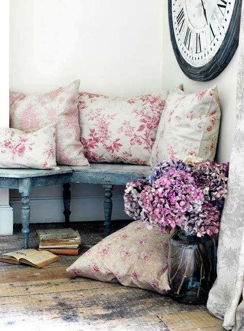 A vintage-style interior corner featuring a distressed blue wooden bench adorned with pink and cream floral pillows, a glass vase holding purple and pink hydrangeas, old books on the wooden floor, and a large antique clock on the wall.