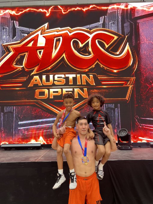 A shirtless father proudly holds his two young sons on his shoulders, all wearing medals, in front of the ADCC Austin Open backdrop with its fiery red design. The scene captures a celebratory moment of accomplishment, reflecting the post's message about cherishing activities with his kids and being proud of their heart and grit during the competition.