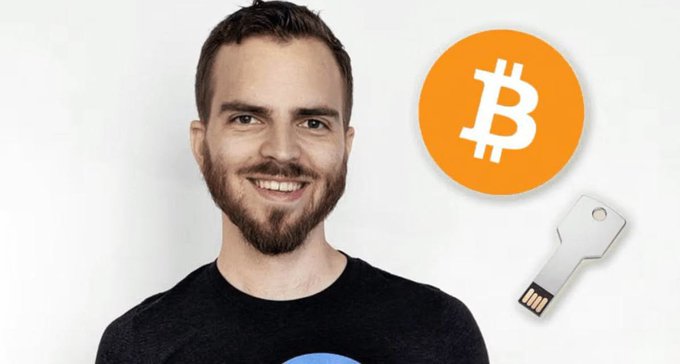 The image shows a smiling man with a beard wearing a black t-shirt, positioned next to an orange Bitcoin logo and a silver IronKey encrypted USB drive. This visually represents Stefan Thomas, the programmer who received 7,002 BTC for creating the "What is Bitcoin?" explainer video in 2011, but lost the password to his IronKey USB—where the Bitcoin is stored—with only 10 attempts remaining before the drive is permanently locked.