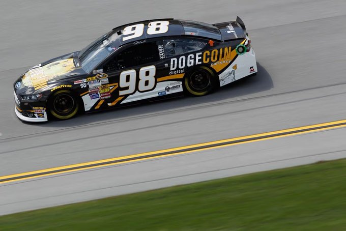 A black #98 NASCAR race car featuring 'DOGECOIN' sponsorship with 'digital currency' text, racing on a track with motion blur visible in the background.