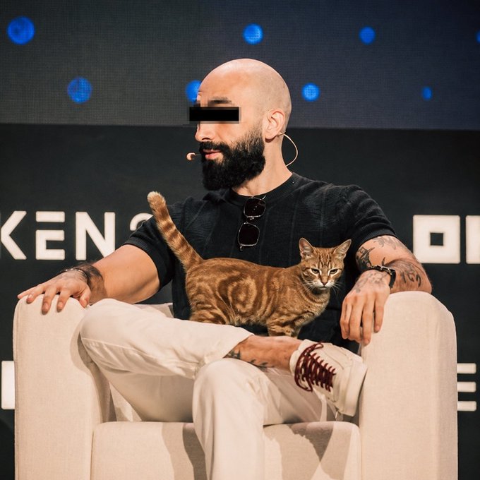 A bald, bearded man sits on a stage with a brown tabby cat resting across his lap, with the partial text "KEN" and "OR" visible on the backdrop behind him. The post references "@mert abi" and uses a cat emoji along with a cryptocurrency symbol, playfully linking the cat in the photo to the "$◎" token mentioned in the message.