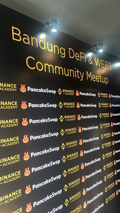 The image shows a large black wall with white text, advertising an event for the "Bandung DeFi & Web3" community meeting at PancakeSwap. The text includes logos of PancakeSwap and BinanceAcademy, indicating that this is likely an event related to cryptocurrency or blockchain technology.
