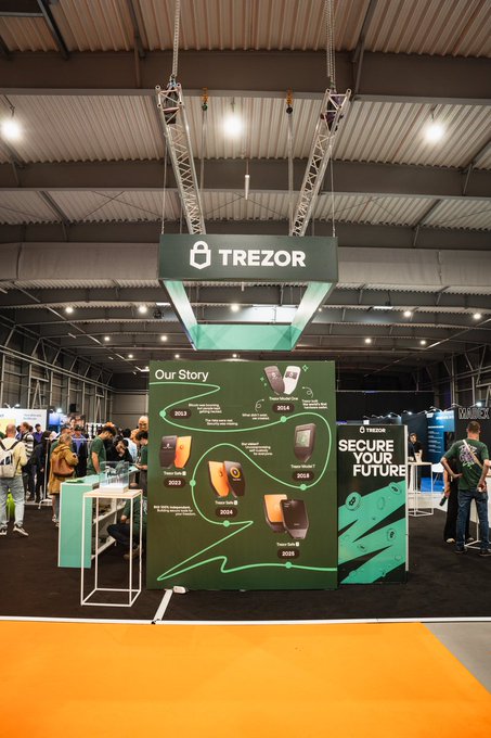 The image shows a trade show booth for Trezor hardware wallets set up inside a building with a high ceiling and white walls. The booth displays various Trezor products, such as smart keys and Bitcoin wallets. A large sign at the front of the booth states that it's an exhibit of inventors of the hardware wallet designed to secure crypto for 2+ million users worldwide. Additionally, there are two people visible in the scene, one standing near a table and another person with a backpack closer to the booth. The image focuses on showcasing the Trezor products at the trade show event.