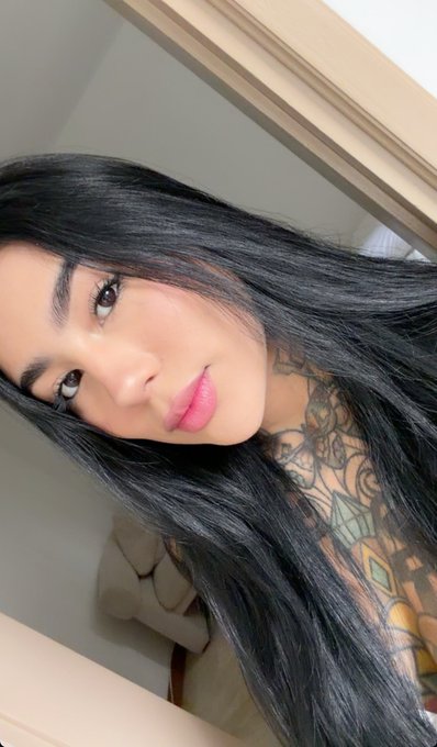 A young woman with long black hair and pink lipstick takes a selfie, showing a large floral and diamond tattoo on her neck and chest.