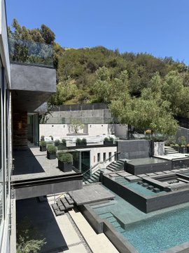 The image shows a large, modern house with an outdoor pool area featuring a glass-enclosed swimming pool surrounded by several lounge chairs. A stone wall can be seen behind the pool, providing a natural border for the space. The house has a balcony overlooking the pool and is situated on a hillside, offering a scenic view of the surrounding landscape.

The post in the image reads "Showings", which suggests that this property might be part of an open house event or a real estate listing showcasing the house's features and layout.