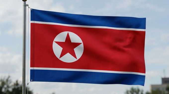 The flag of North Korea waves against a partly cloudy sky, featuring its iconic red star inside a white circle on a wide red stripe, bordered by blue and thin white stripes.