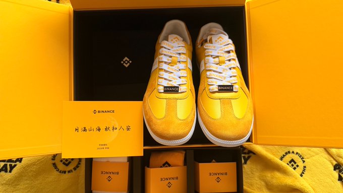 A pair of yellow Binance branded sneakers displayed in their signature yellow box with a Chinese calligraphy card.