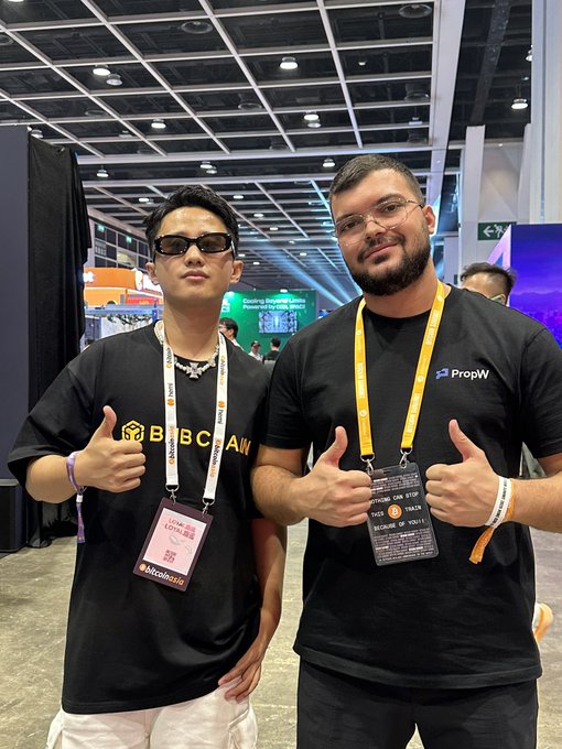 Two men pose for a photo at what appears to be a crypto/blockchain conference, both giving thumbs up. The man on the left wears a black "Boba Chain" t-shirt with a "LUNA LOTTERY 5" lanyard, while the man on the right wears a "PropW" shirt and a "NEXT BLOCK Asia #10" conference badge, capturing a chance reunion between former colleagues at a Bitcoin/AI event as referenced in the post.