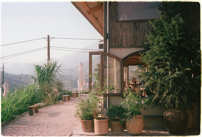 The image shows a house with potted plants placed outside near a path leading up to an open door. The scene also includes a bench and a tree nearby. There are two people standing close together, possibly engaged in conversation or admiring their surroundings. A person is sitting on one of the benches, while another individual stands next to them. In addition, there is a potted plant placed near the path, adding greenery to the scene. The image also contains text that reads "Good afternoon everyone Quick thought for your afternoon. We lock our doors. We use passwords. We enable 2FA. But our crypto wallets? Still vulnerable to what’s coming next."