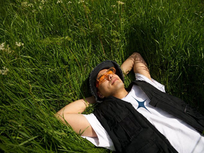 A person wearing orange-tinted sunglasses and a dark hat relaxes on their back in lush green grass with hands behind their head.