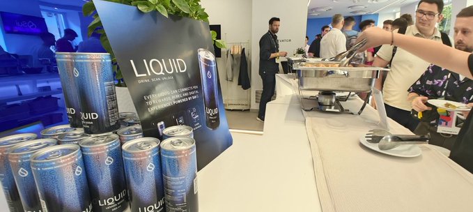 Cans of "LIQUID" sparkling water are displayed at SuiHub Athens, a blockchain-enabled product featured at the crypto hub's event. The promotional sign highlights its key features: "DRINK. SCAN. ENJOY," "JUST PLUG A WALLET," "100% FLAVORS," and "SUPERCHARGED POWERED BY SUI," illustrating a real-world application of blockchain technology.