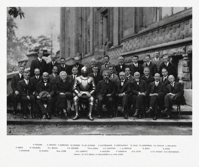 A black-and-white historical photograph of a group of distinguished men, including a knight in armor at the center, identified by caption text as attendees of the Fifth Solvay Conference including Einstein, Bohr, Marie Curie, Schrödinger, and Heisenberg.