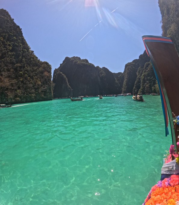 A bright, sunlit view of turquoise water surrounded by dramatic limestone karst cliffs, with traditional longtail boats floating on the surface, likely Maya Bay or a similar location in Thailand.