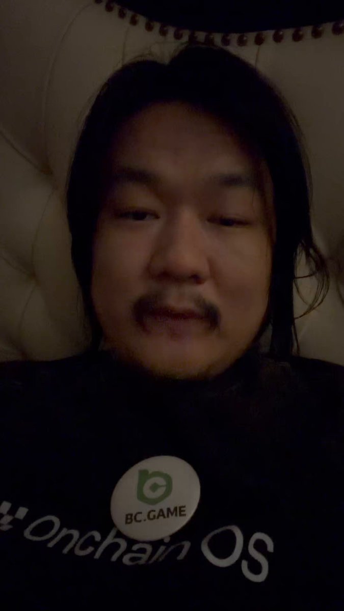 The image features a man wearing a black shirt with a green "BC Game" logo on it. He has long hair and appears to be looking directly at the camera while sitting or lying down, possibly taking a selfie. The background of the photo is blurred, but there are some chairs visible in the scene.