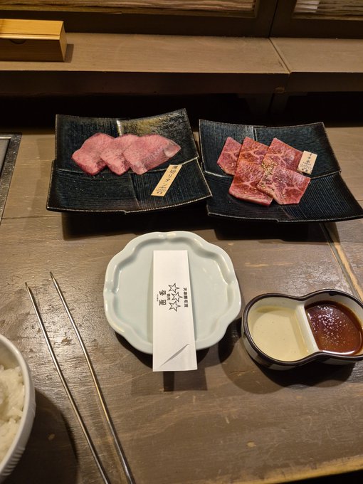 Photo of a Japanese meal featuring two plates of premium raw beef slices, a small dish with sauces (white and red), chopsticks, and a wrapped item on a wooden counter.