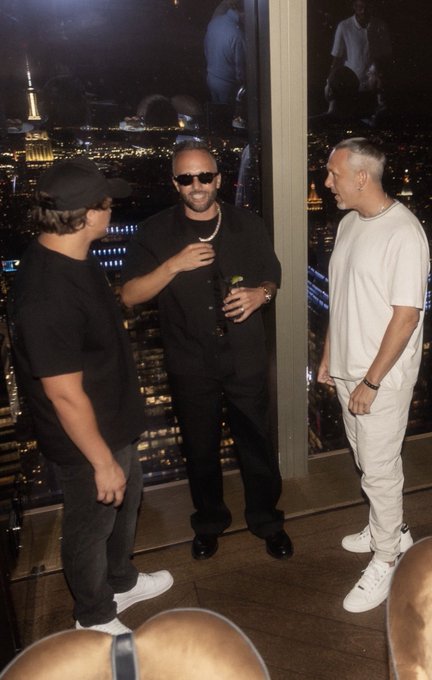 Three men dressed in black and white casual attire standing in conversation at what appears to be a high-rise venue with city lights visible through the windows behind them.