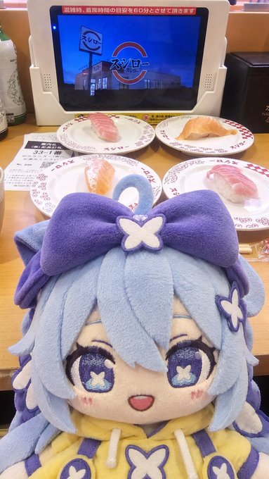 A blue-haired plush doll with a large purple bow sits in front of a conveyor belt sushi restaurant's tablet screen displaying 'スタート' (Start), with plates of sushi visible behind it.