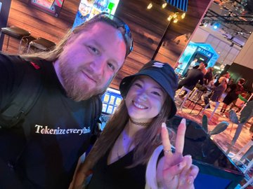 A selfie of a smiling couple in what appears to be a bar or restaurant; the man has dreadlocks and wears a 'Tokenize everything' shirt, while the woman wears a black bucket hat and flashes a peace sign.