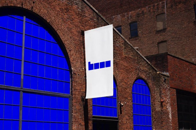 A white vertical banner hanging on a brick building with blue arched windows, displaying a blue geometric logo resembling the letters 'L' and 'H'.